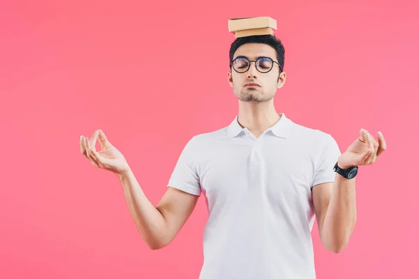 Handsome student with closed eyes meditating with books on head isolated on pink — Stock Photo
