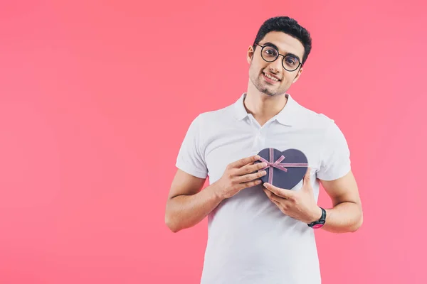 Sorrindo bonito homem segurando caixa de presente em forma de coração perto do coração isolado em rosa, conceito de dia dos namorados — Fotografia de Stock