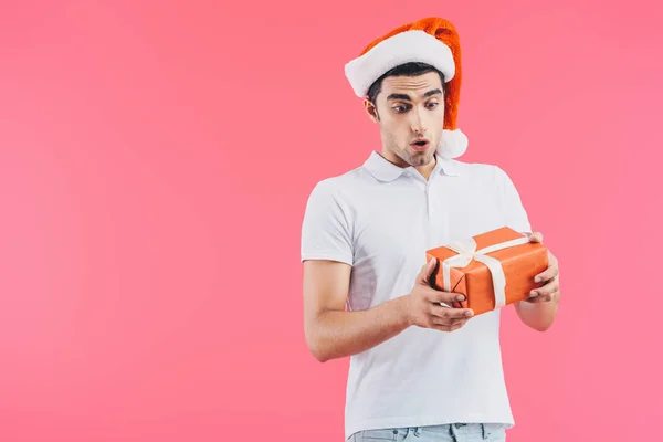 Shocked handsome man in santa hat looking at gift box isolated on pink, new year concept — Stock Photo