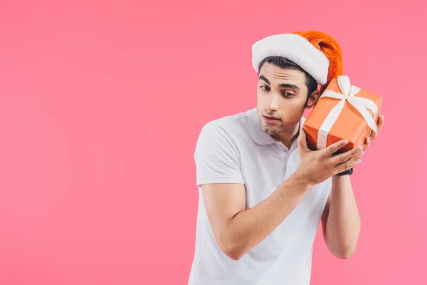 Aufgeregter gutaussehender Mann mit Weihnachtsmannhut hört Geschenkbox isoliert auf rosa, Neujahrskonzept — Stockfoto