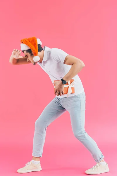 Vista laterale dell'uomo in cappello di Babbo Natale contenente scatola regalo e volto di copertura isolato su rosa, concetto di nuovo anno — Foto stock