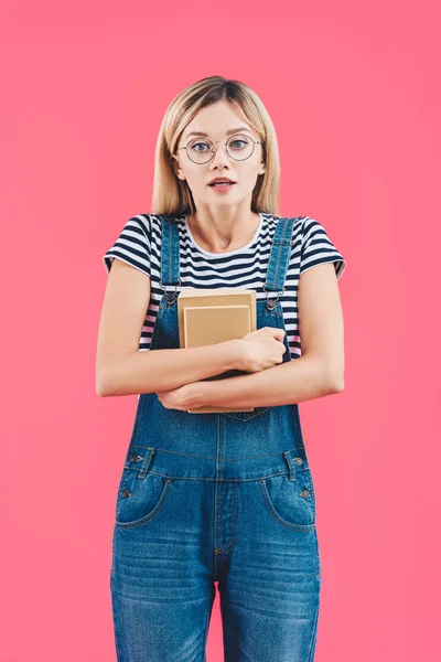 Scared student in striped shirt and denim overall holding books and looking at camera isolated on pink — Stock Photo