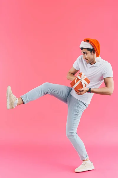 Grimacing handsome man in santa hat and raised leg holding gift box isolated on pink, new year concept — Stock Photo