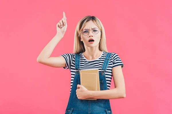 Ritratto di giovane studente in occhiali con libri che puntano in alto isolato su rosa — Foto stock