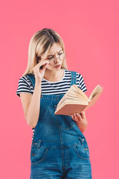 Retrato de mujer joven en anteojos libro de lectura aislado en rosa - foto de stock