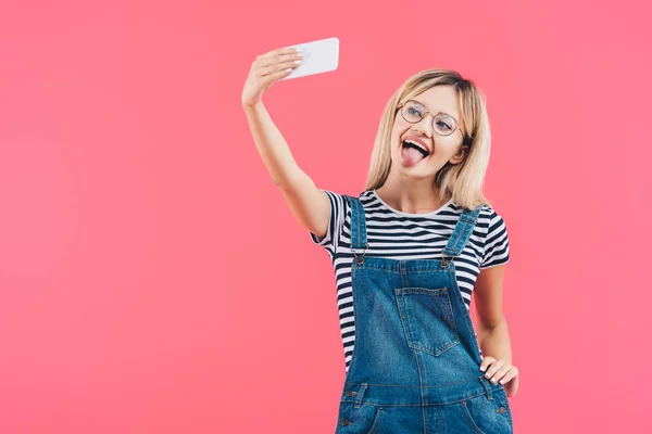 Retrato de mulher colando a língua para fora ao tomar selfie no smartphone isolado no rosa — Fotografia de Stock