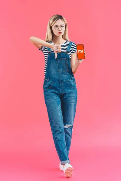 Mujer joven con teléfono inteligente con el logotipo de xiaomi que muestra el pulgar hacia abajo en el fondo rosa - foto de stock