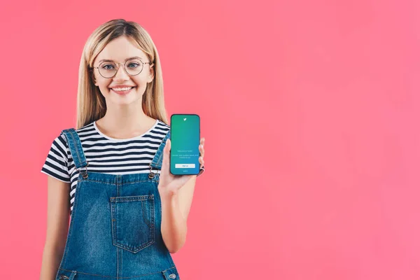 Portrait of smiling woman in eyeglasses showing smartphone with twitter logo isolated on pink — Stock Photo
