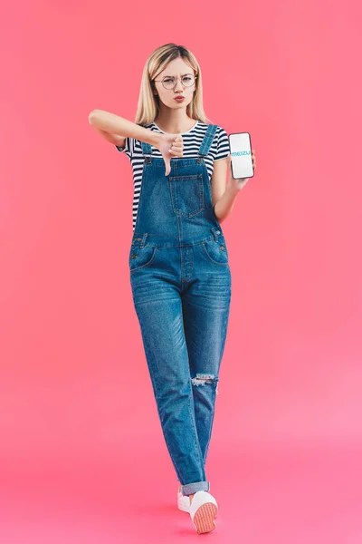 Young woman with smartphone with meizu logo showing thumb down on pink backdrop — Stock Photo