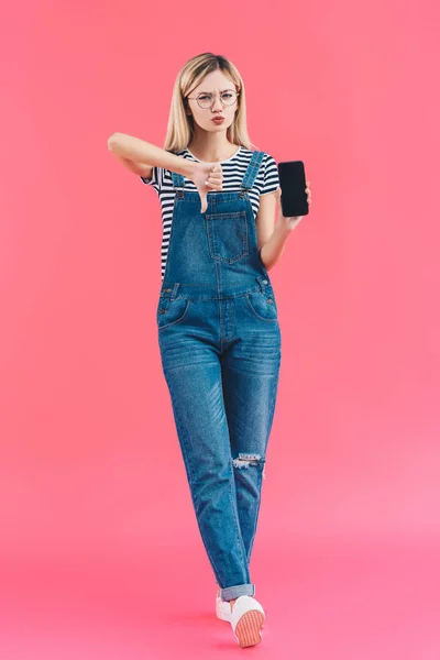 Mujer joven con teléfono inteligente con pantalla en blanco que muestra el pulgar hacia abajo en el fondo rosa - foto de stock