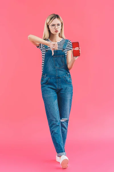 Young woman with smartphone with youtube logo showing thumb down on pink backdrop — Stock Photo