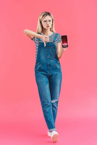 Mujer joven con teléfono inteligente que muestra el pulgar hacia abajo en el fondo rosa - foto de stock
