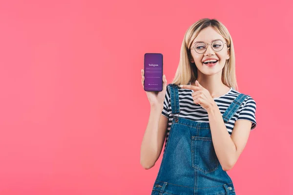 Porträt einer lächelnden Frau mit Brille, die auf ihr Smartphone zeigt, mit Instagram-Zeichen auf rosa — Stockfoto