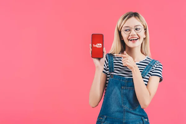 Ritratto di donna sorridente in occhiali che punta verso smartphone con segno youtube isolato su rosa — Foto stock