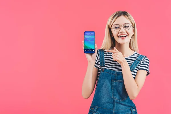 Retrato de mujer sonriente en gafas apuntando al teléfono inteligente con signo de reserva aislado en rosa - foto de stock