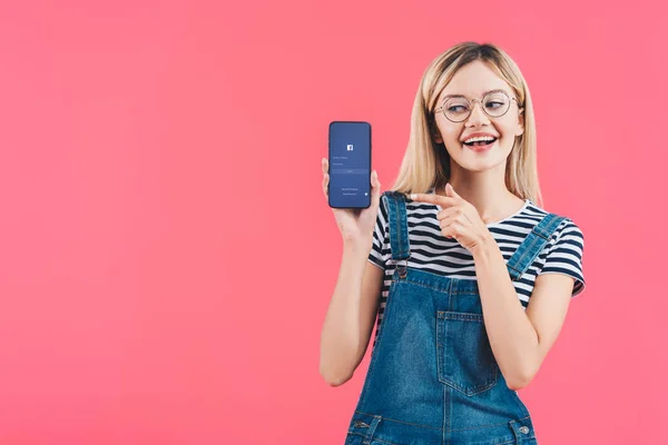 Portrait of smiling woman in eyeglasses pointing at smartphone with facebook logo isolated on pink — Stock Photo