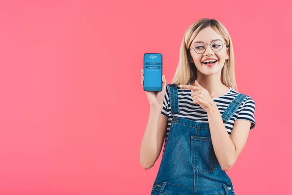 Portrait of smiling woman in eyeglasses pointing at smartphone with skype logo isolated on pink — Stock Photo