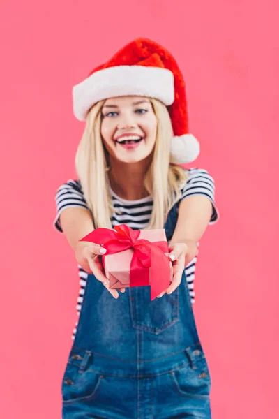 Ritratto di giovane donna allegra in cappello di Babbo Natale in possesso di regalo isolato su rosa — Foto stock