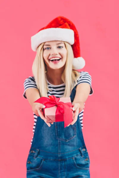 Portrait de jeune femme gaie en santa claus chapeau tenant cadeau isolé sur rose — Photo de stock