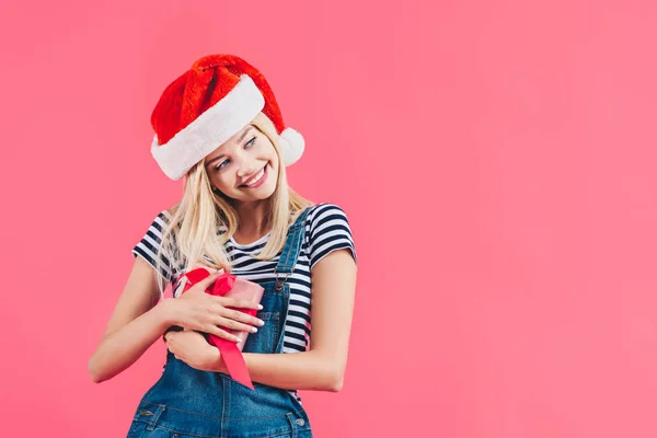 Ritratto di giovane donna felice in cappello di Babbo Natale in possesso di regalo isolato su rosa — Foto stock