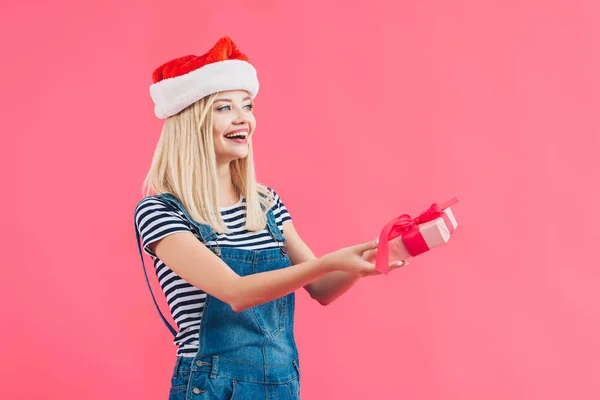 Giovane donna sorridente in cappello di Babbo Natale in possesso di regalo isolato su rosa — Foto stock