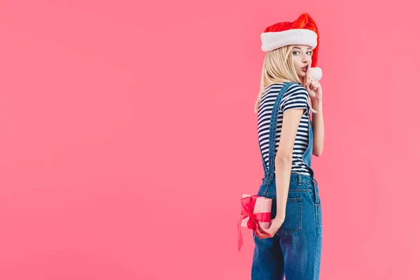 Vista laterale della donna in cappello Babbo Natale con regalo che mostra segno di silenzio isolato su rosa — Foto stock