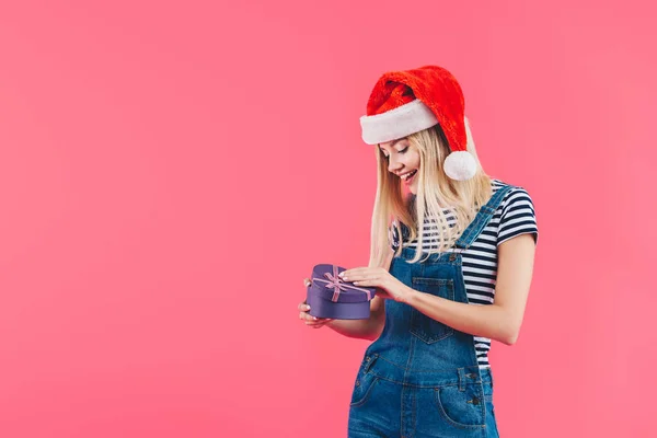 Sorridente giovane donna in cappello Babbo Natale con regalo a forma di cuore isolato su rosa — Foto stock