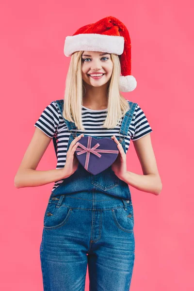 Портрет улыбающейся женщины в шляпе Санта-Клауса с подарком в форме сердца, изолированным на розовом — стоковое фото