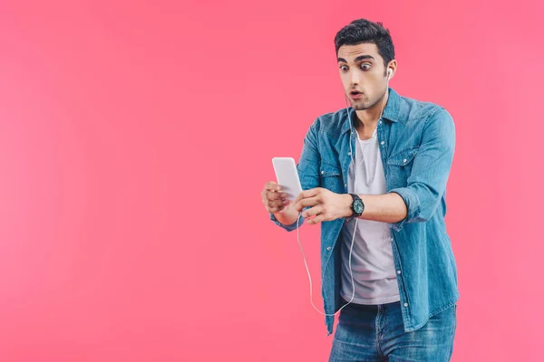 Портрет шокованого чоловіка в навушниках, який дивиться на смартфон в руках ізольовано на рожевому — стокове фото