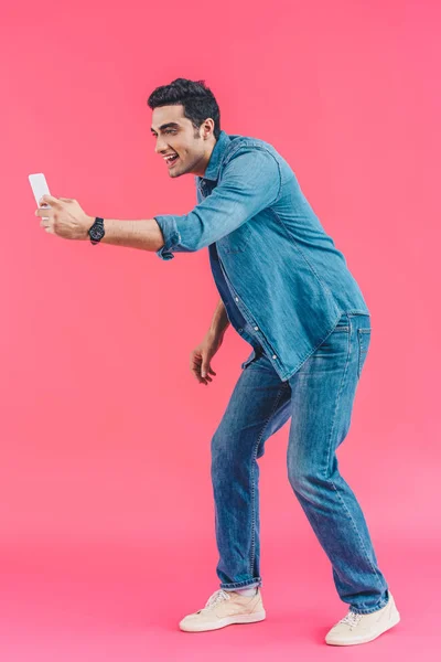 Vista lateral del hombre alegre tomando una foto en el teléfono inteligente en el telón de fondo rosa - foto de stock