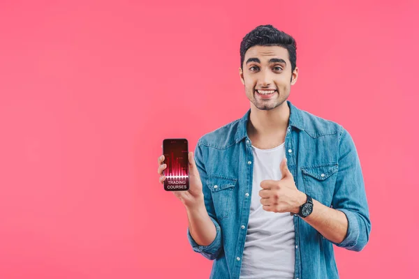 Lächelnder junger Mann macht Daumen-hoch-Geste und zeigt Smartphone mit Handelskursen isoliert auf rosa — Stockfoto