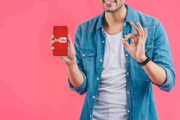 Partial view of young man doing ok sign and showing smartphone with youtube website isolated on pink — Stock Photo