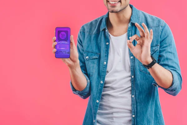 Vista parcial de un joven haciendo signo de ok y mostrando el teléfono inteligente con compras aisladas en rosa - foto de stock