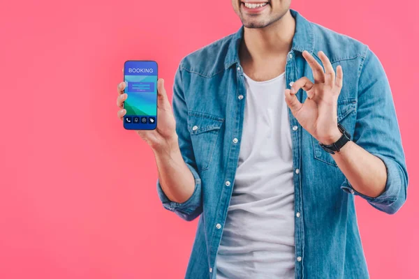 Vista parcial de un joven haciendo signo de ok y mostrando el teléfono inteligente con reserva aislada en rosa - foto de stock