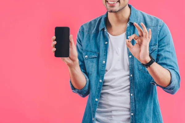 Vista parcial del joven haciendo signo de ok y mostrando el teléfono inteligente con pantalla en blanco aislado en rosa - foto de stock