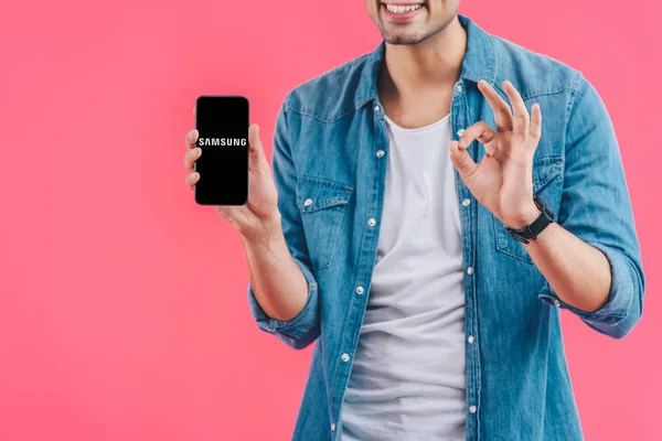 Vista parcial de hombre joven haciendo signo de ok y mostrando Samsung teléfono inteligente aislado en rosa - foto de stock