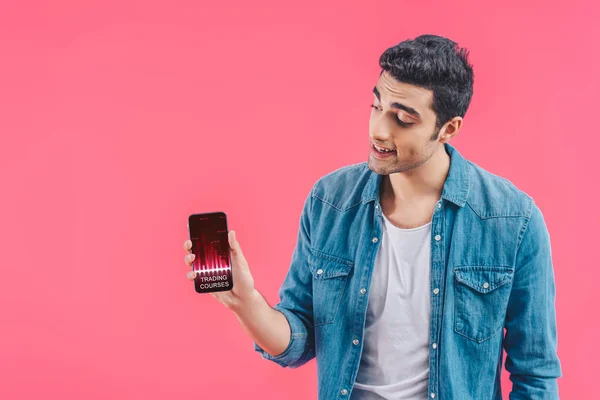 Lächelnder junger Mann zeigt Smartphone mit Handelskursen isoliert auf rosa — Stockfoto