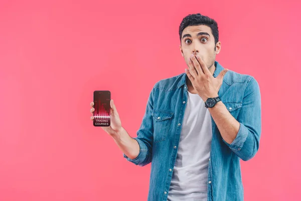 Schockierter junger Mann verdeckt Mund mit der Hand und zeigt Smartphone mit Handelskursen isoliert auf rosa — Stockfoto