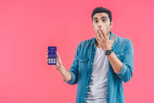 Sorprendido joven cubriendo boca a mano y mostrando teléfono inteligente con aplicación médica aislado en rosa - foto de stock