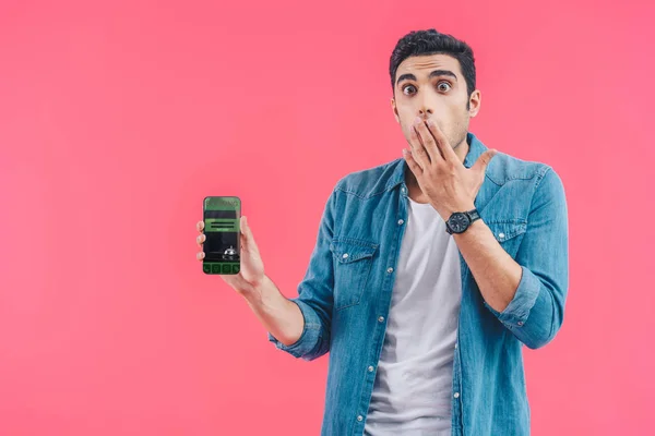 Schockierter junger Mann verdeckt Mund mit der Hand und zeigt Smartphone mit Buchung isoliert auf rosa — Stockfoto