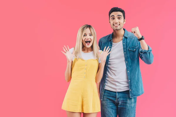 Excitada pareja joven haciendo gestos con las manos y mirando a la cámara aislada en rosa - foto de stock