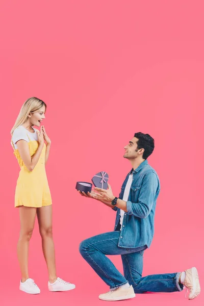 Вид сбоку мужчины на согнутом колене, делающего предложение о браке удивленной подруге, жестикулирующей руками, изолированными от розового — стоковое фото