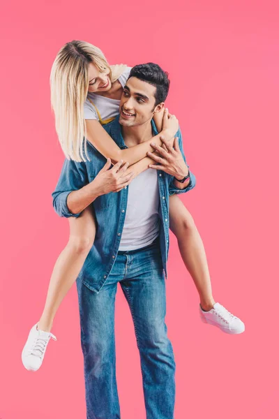 Handsome young man doing piggyback ride to smiling girlfriend isolated on pink — Stock Photo