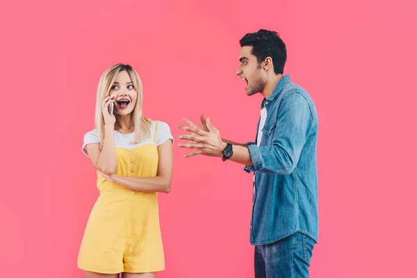 Side view of aggressive man gesturing by hand and yelling at girlfriend while she talking on smartphone isolated on pink — Stock Photo