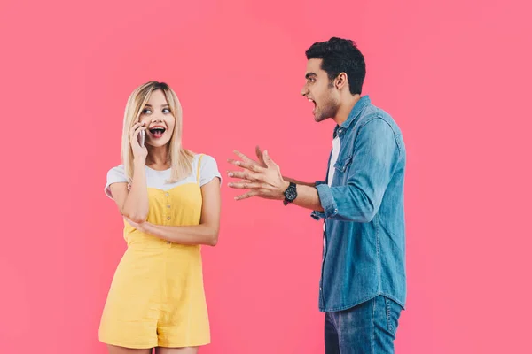 Vista laterale di uomo aggressivo gesticolando a mano e urlando alla fidanzata mentre lei parla su smartphone isolato su rosa — Foto stock