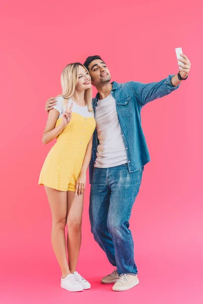 Красивий молодий чоловік бере селфі з дівчиною робить знак миру на смартфоні ізольовано на рожевому — стокове фото