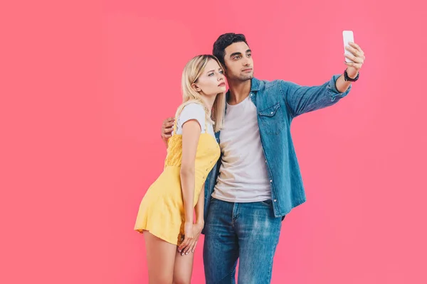 Serious young couple taking selfie on smartphone isolated on pink — Stock Photo