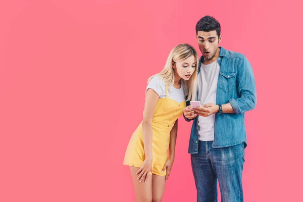 Sorprendido joven mirando el teléfono inteligente mientras que su novia de pie cerca aislado en rosa - foto de stock