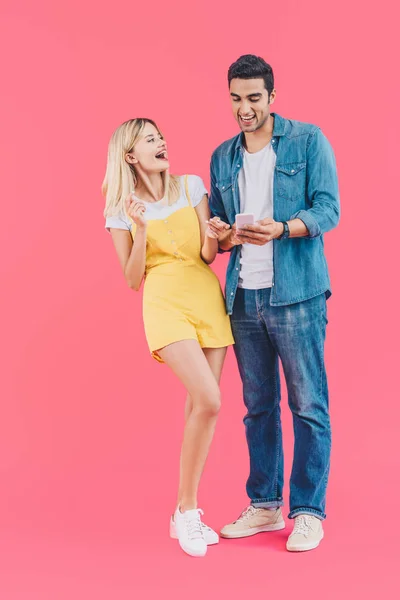 Cheerful young man showing smartphone to laughing girlfriend isolated on pink — Stock Photo