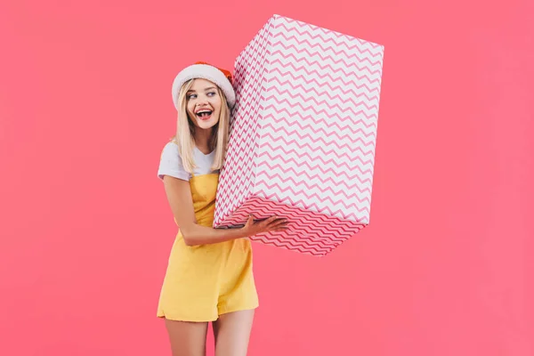 Cheerful young woman in christmas hat listening big gift box isolated on pink — Stock Photo